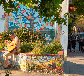 An older man in Fairfax California sitting on a planters edge playing his guitar in front of an expressive colorful mural.